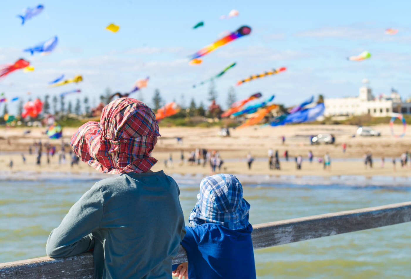 Two young children wearing hats looking out over the water to a kite festival on the beach
