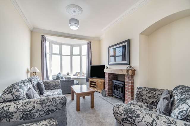 Bright living room with patterned grey sofas, a wooden coffee table, a brick fireplace with a stove, and a large bay window with grey curtains letting in natural light.
