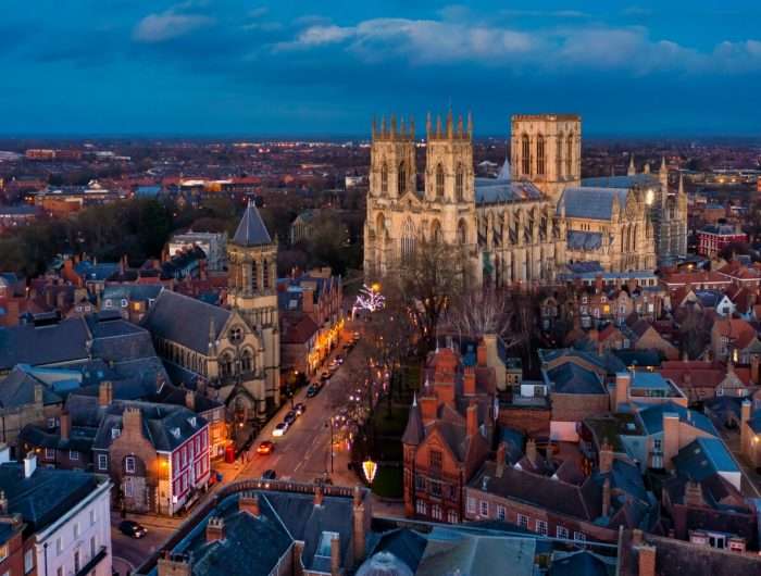 A twilight aerial view of York, England, featuring the illuminated York Minster cathedral towering above the historic red-brick buildings and narrow streets below, with city lights glowing into the distance under a deep blue sky.