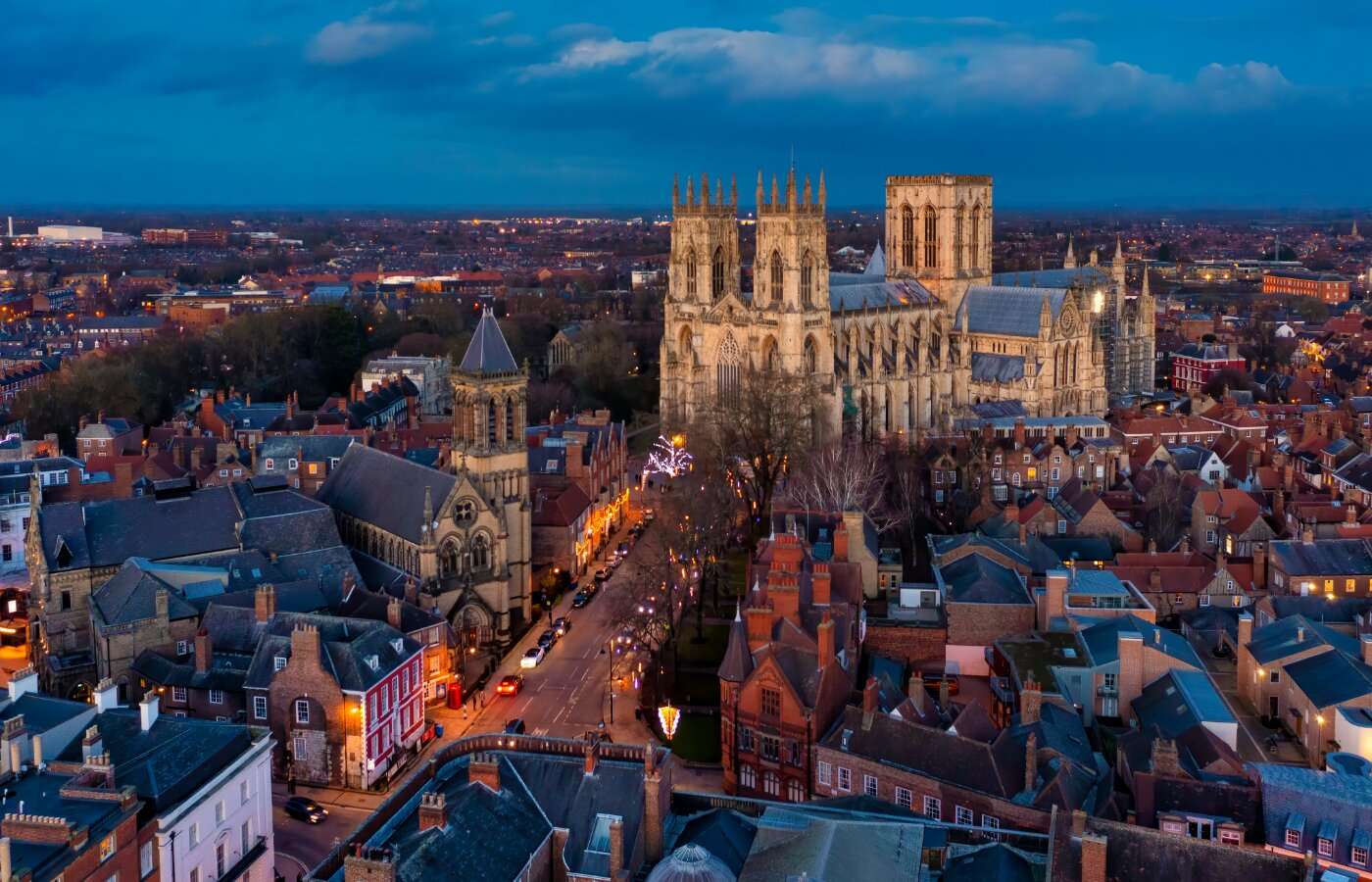 A twilight aerial view of York, England, featuring the illuminated York Minster cathedral towering above the historic red-brick buildings and narrow streets below, with city lights glowing into the distance under a deep blue sky.