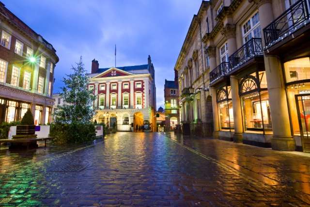 A wet cobblestone street in York at dusk, reflecting colorful lights from surrounding historic buildings, leading toward the grand façade of York’s Guildhall with a Christmas tree in the square.