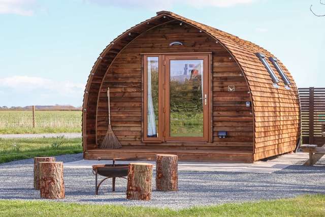 Wooden glamping pod with curved timber cladding, a glass front door, and a small patio featuring log stools and a fire pit, set in an open rural landscape.