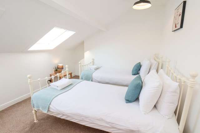 Bright attic bedroom with two white metal-frame single beds, soft teal cushions, a skylight window, and a small bedside table with a lamp and plant.