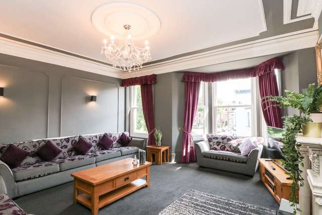 Elegant living room with grey sofas, purple cushions and curtains, a wooden coffee table, and a chandelier, featuring a large bay window that fills the space with natural light.