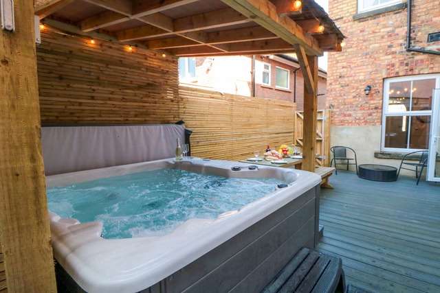 Outdoor deck area with a bubbling hot tub under a wooden pergola, surrounded by privacy fencing, with seating and a table set for drinks and snacks.