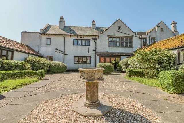 Historic manor-style building with white rendered walls, gabled roofs, and tall chimneys, overlooking a central courtyard garden with trimmed hedges and a stone pedestal feature.