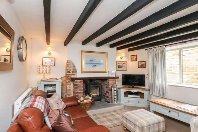 Cosy living room with exposed wooden beams, a brown leather sofa, a plaid footstool, and a brick fireplace with a wood-burning stove, decorated with coastal artwork and soft lighting.