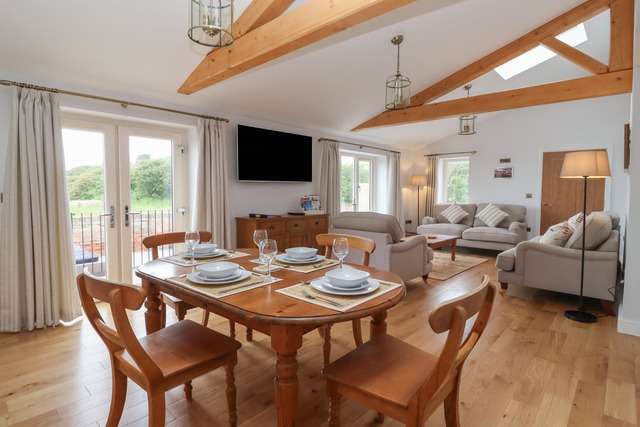 Spacious open-plan dining and living area with wooden beams, a wooden dining table set for four, light grey sofas, and large windows and doors overlooking greenery.